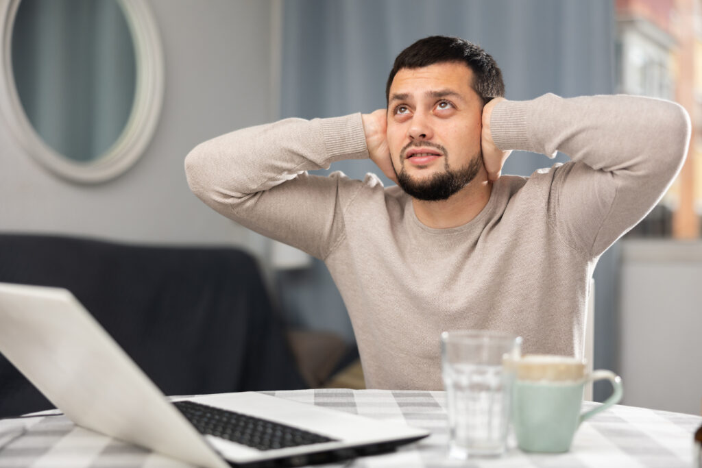 Annoyed unhappy bearded guy working at home at table with laptop, covering ears with his hands because of loud noise in neighbors apartment from above