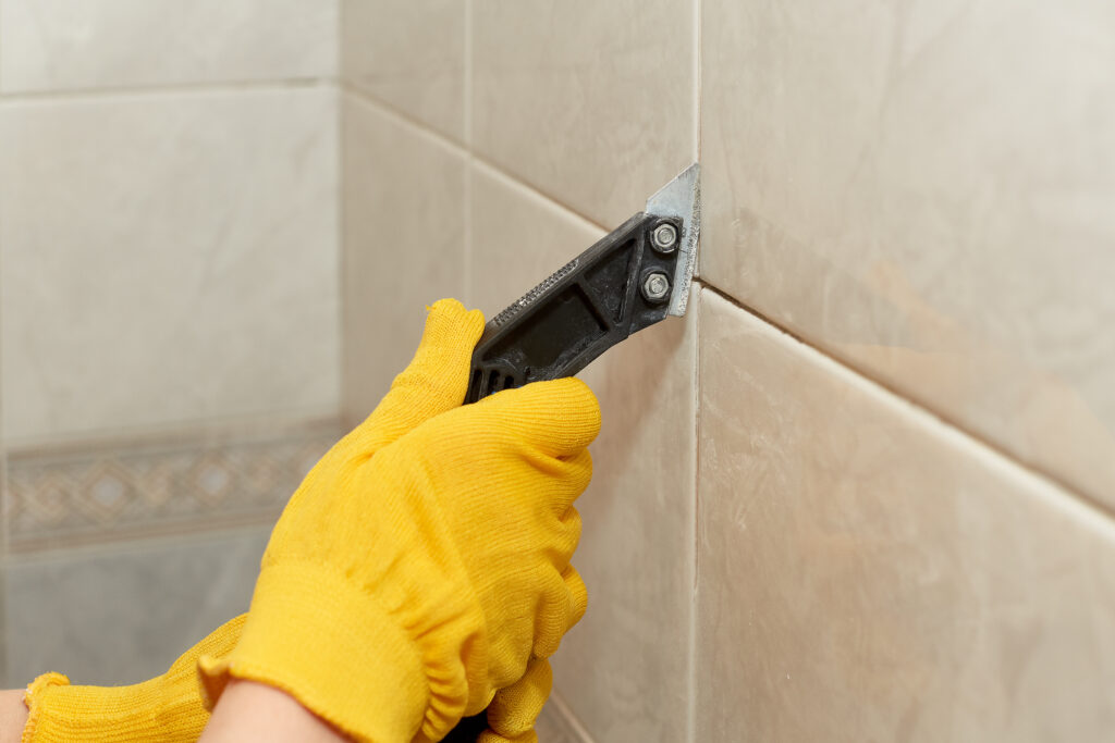 Male hands with knife removing the old grout. Replacing old grout between tiles. Raking out tiles for regrouting.