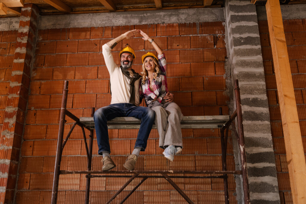Two individuals are sitting on a scaffolding in a building that is being built. They wear yellow hard hats and are smiling as they pose for the camera. It is daytime.