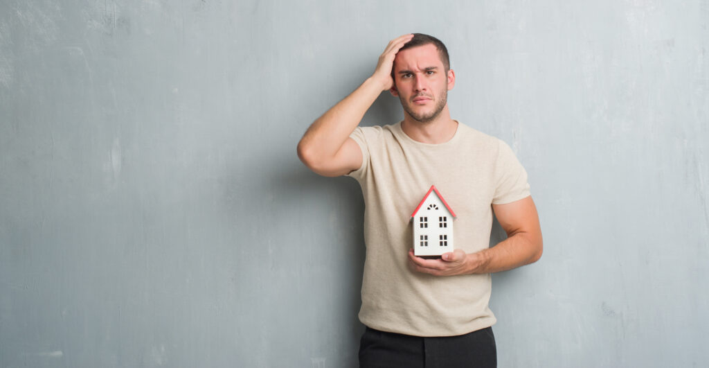 Young caucasian real state agent man over grey grunge wall holding house stressed with hand on head, shocked with shame and surprise face, angry and frustrated. Fear and upset for mistake.