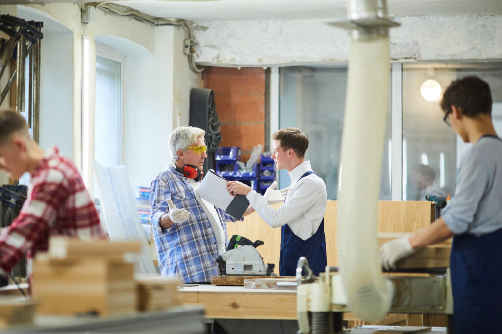 Serious strict young manager poking senior employee with clipboard and scolding him for mistakes in factory workshop