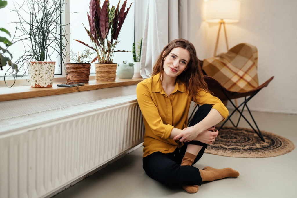 Cute young woman relaxing on the floor near a warm radiator in her socks grinning quietly at the camera with a friendly expression side lit by a large window