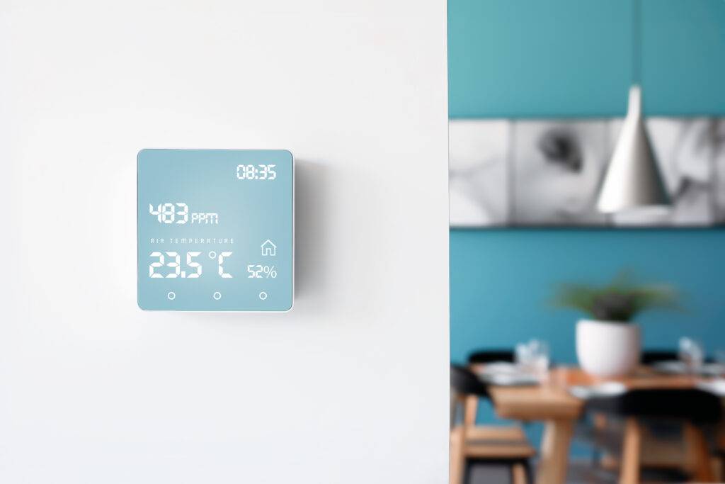 A smart digital thermostat with air quality display on a white wall in a cozy modern living room setting. Advanced home automation for improved everyday comfort and lifestyle. Copy space.